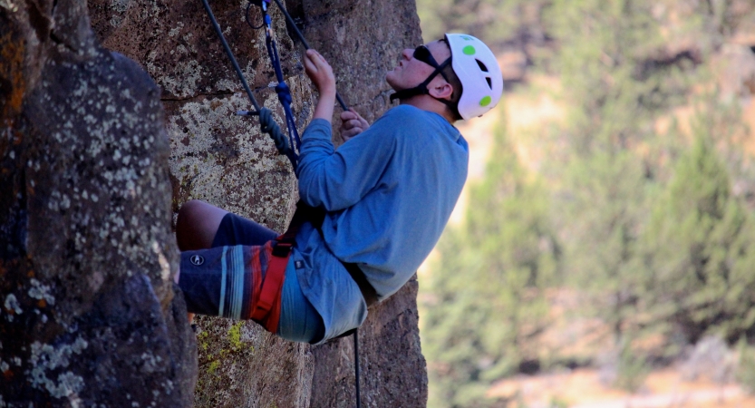 Wearing safety gear and secured by ropes, a person climbs a rock wall. There are trees in the distance in the background. 